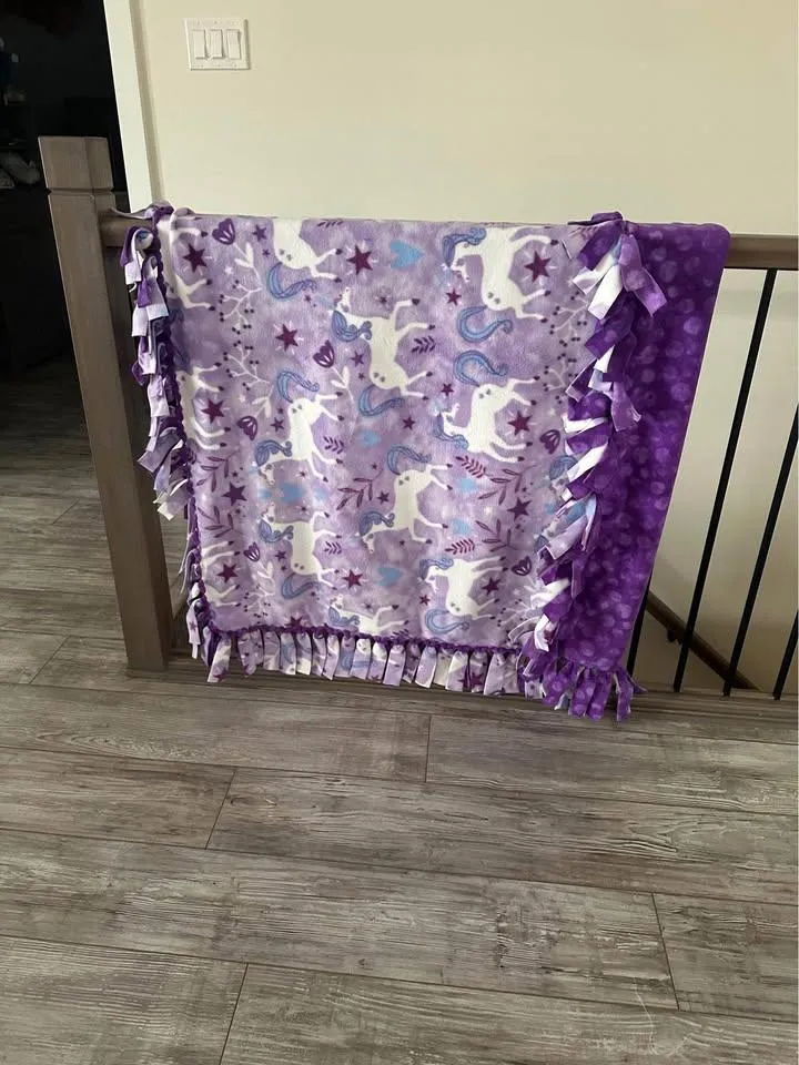 Purple and white unicorn print fleece tie blanket with a knotted fringe draped over a railing